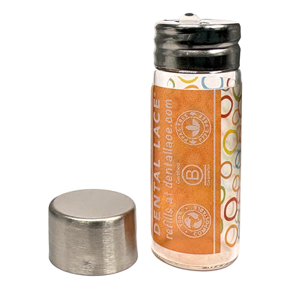Cylindrical glass floss container with a metallic cap and orange ‘Dental Lace’ label on a white background.