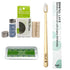 A bamboo toothbrush with a light wood handle and white bristles stands upright next to its white and green branded box. Surrounding it are included accessories: a small glass bottle of dental lace refill, a metal cap, two silk floss spools, and a silver tray with a green Unpaste toothpaste tablet packet.