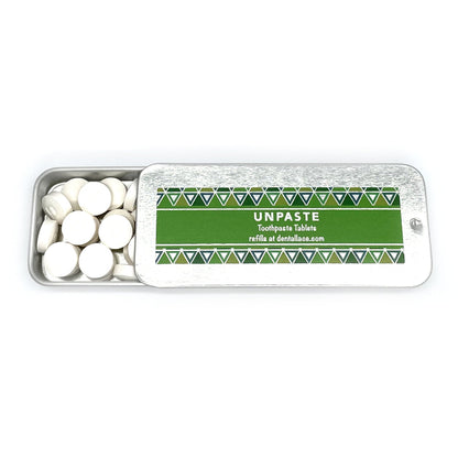 Opened metallic tin with unpaste fluoride free toothpaste tablets and a green design on a white background.